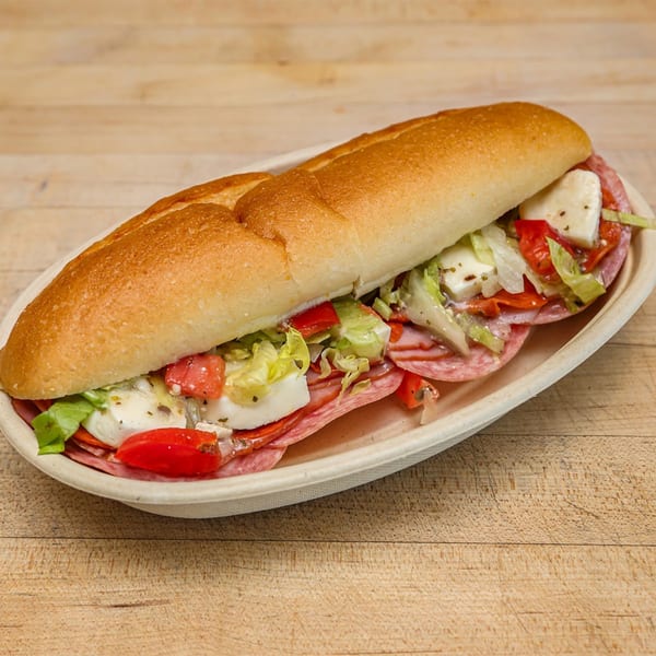 A Roundabout Pizza Italian sub sandwich with salami, mozzarella, lettuce, and tomatoes on a wooden table.